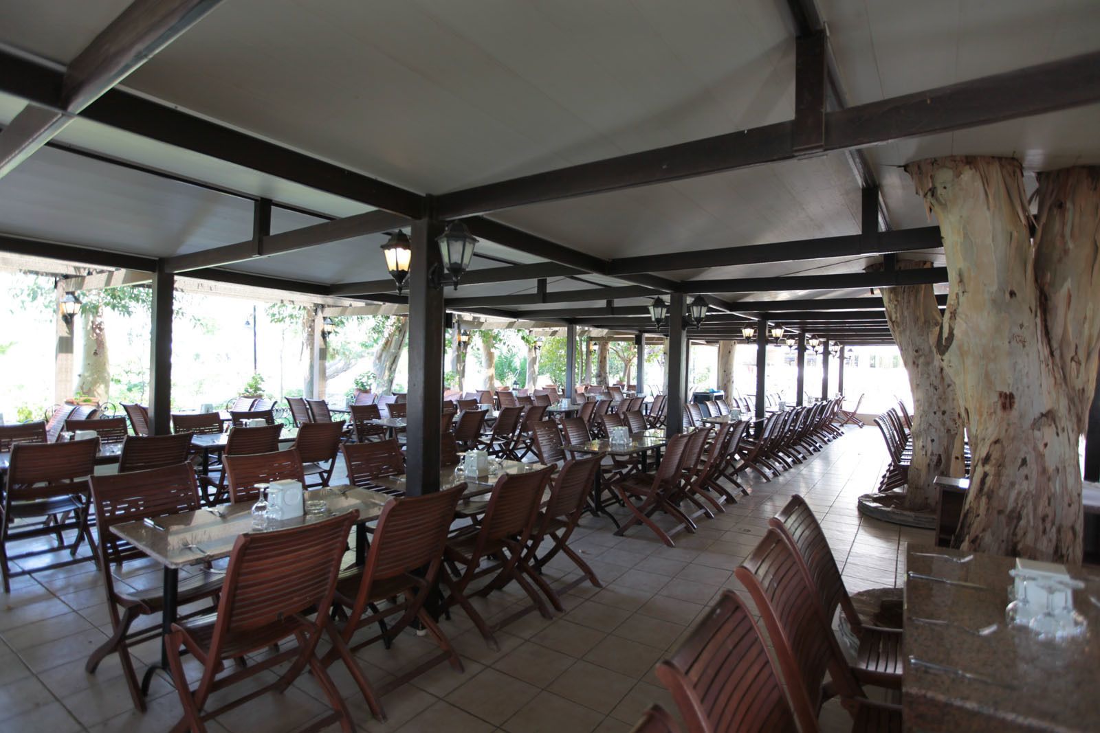 Un espace extérieur abrité, aménagé pour les repas, avec des rangées de tables et de chaises, de grands arbres et un sol carrelé.