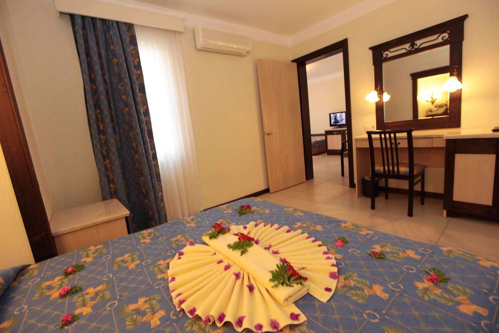 A hotel room with a bed, a desk, a chair, a mirror, and a door leading to another room.