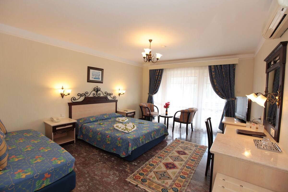 A hotel room with a large bed, two smaller beds, a desk with a chair, a TV, and a chandelier.
