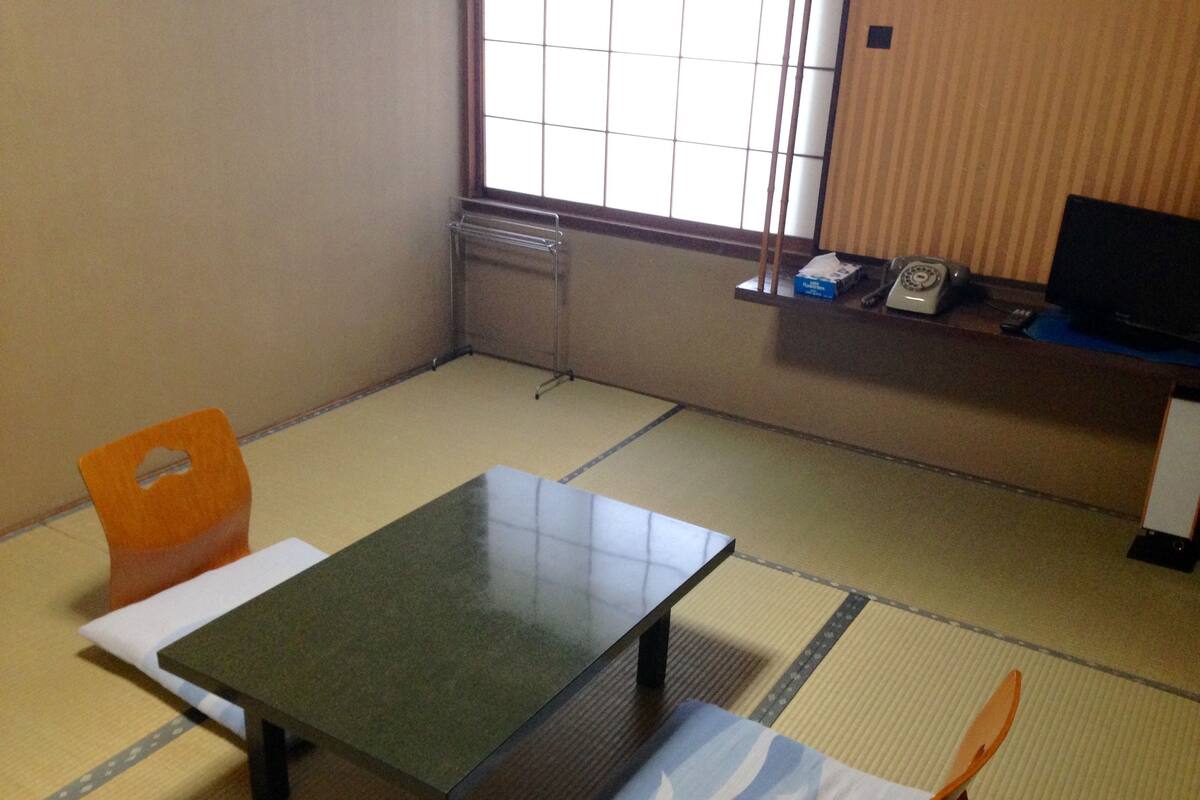 A room with tatami flooring, a low table, a chair, a television on a stand, and a window with blinds.