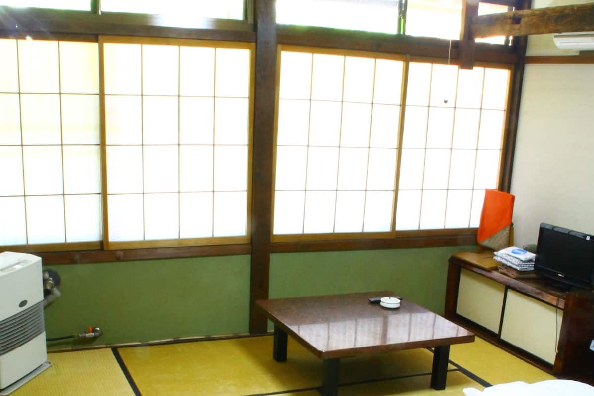 A traditional Japanese room with tatami flooring, a futon bed, a small table with a lamp, and a television on a cabinet.