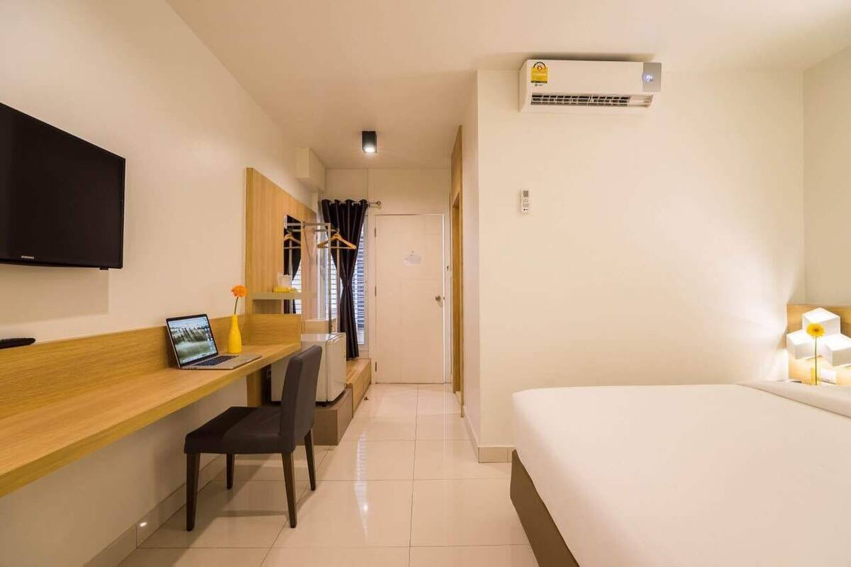 A hotel room with a bed, a desk with a laptop, a TV, and an air conditioning unit.