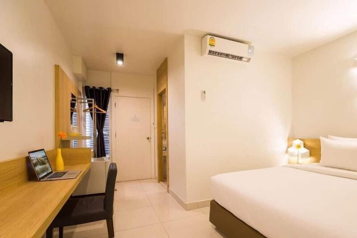 A hotel room with a bed, a desk with a laptop, a chair, an air conditioning unit, and a window with curtains.