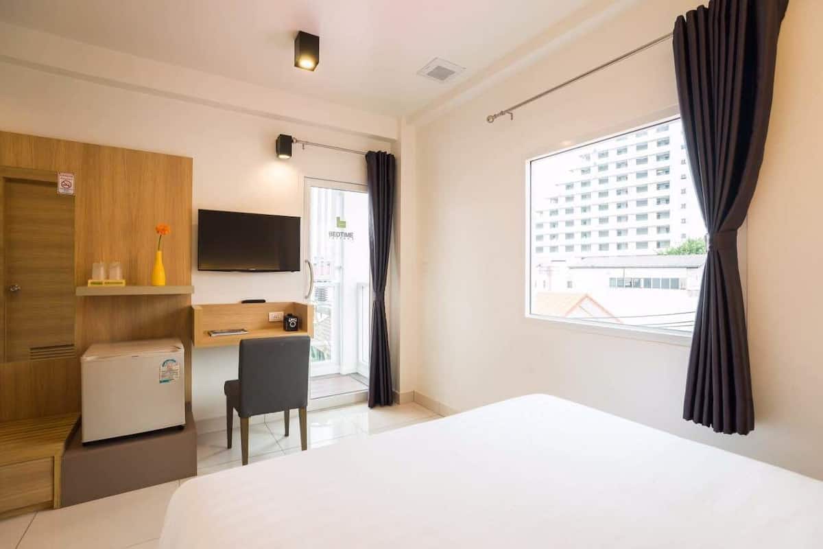 A hotel room with a bed, a desk, a chair, a television, a small refrigerator, and a window with curtains.