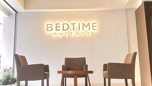 Um interior moderno com um ventilador de teto, uma placa na parede com a inscrição "BEDTIME PATTAYA" e três cadeiras ao redor de uma pequena mesa.
