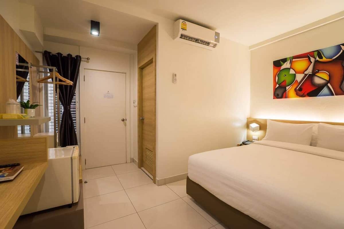 A hotel room with a bed, a desk, a wardrobe, and an air conditioning unit.