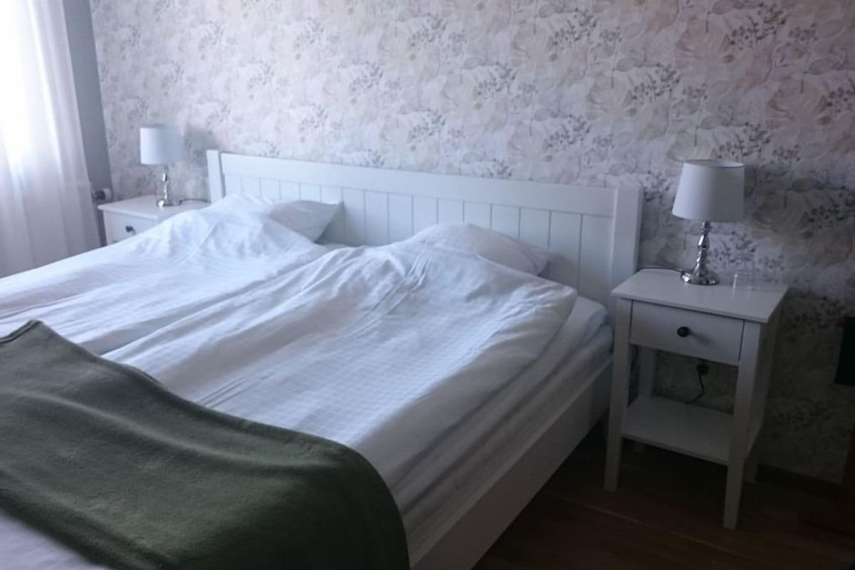 A neatly made bed with white linens and a green throw blanket, a white bedside table with a lamp, and a floral wallpaper pattern on the wall.