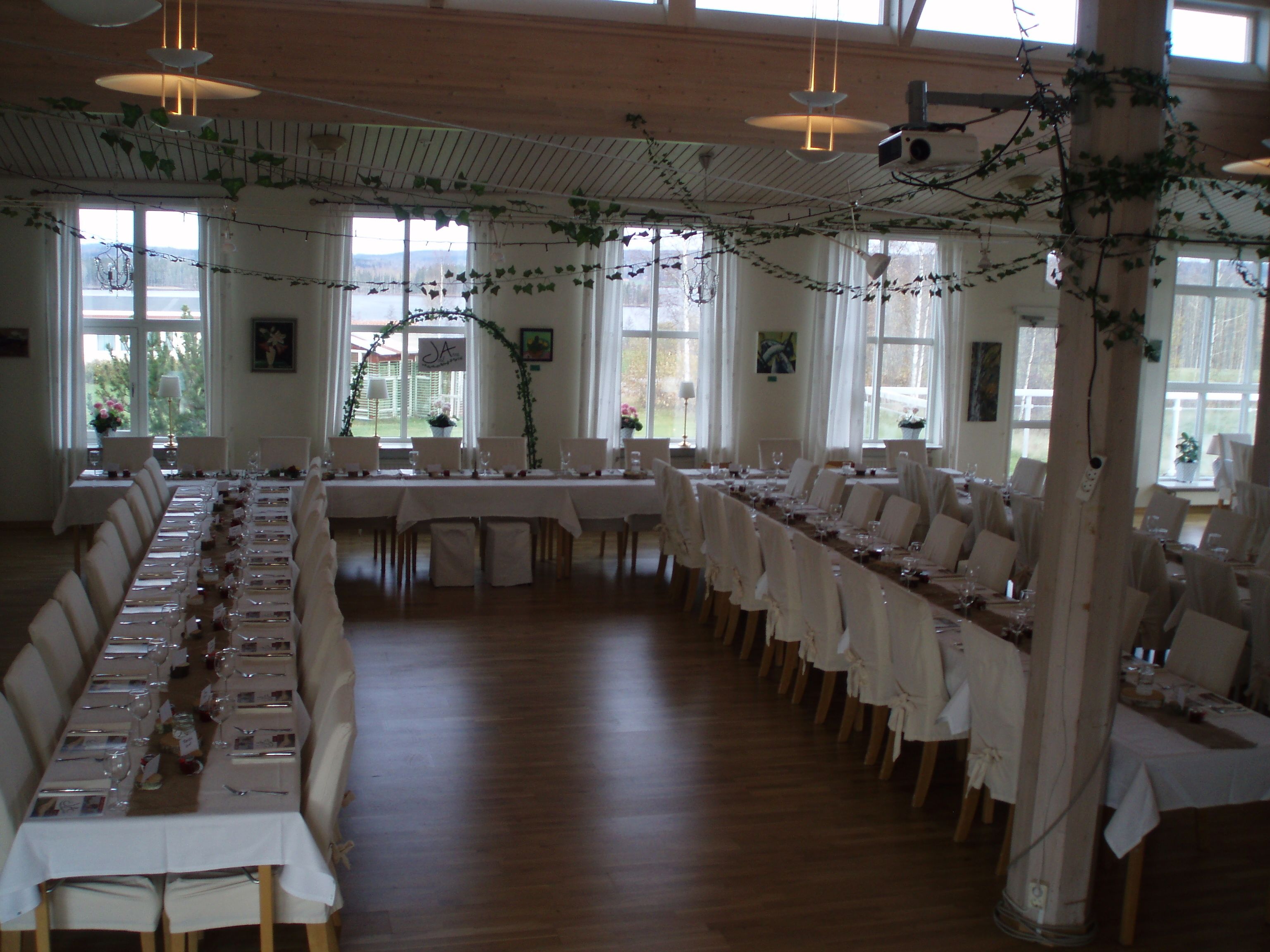 A long dining table set for a formal event with white tablecloths and chairs, large windows with natural light, and decorative greenery.