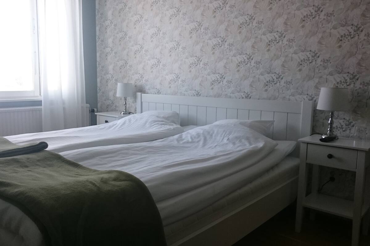 Una cama doble con ropa de cama blanca, una mesita de noche con lámpara y un estampado floral en el papel tapiz.