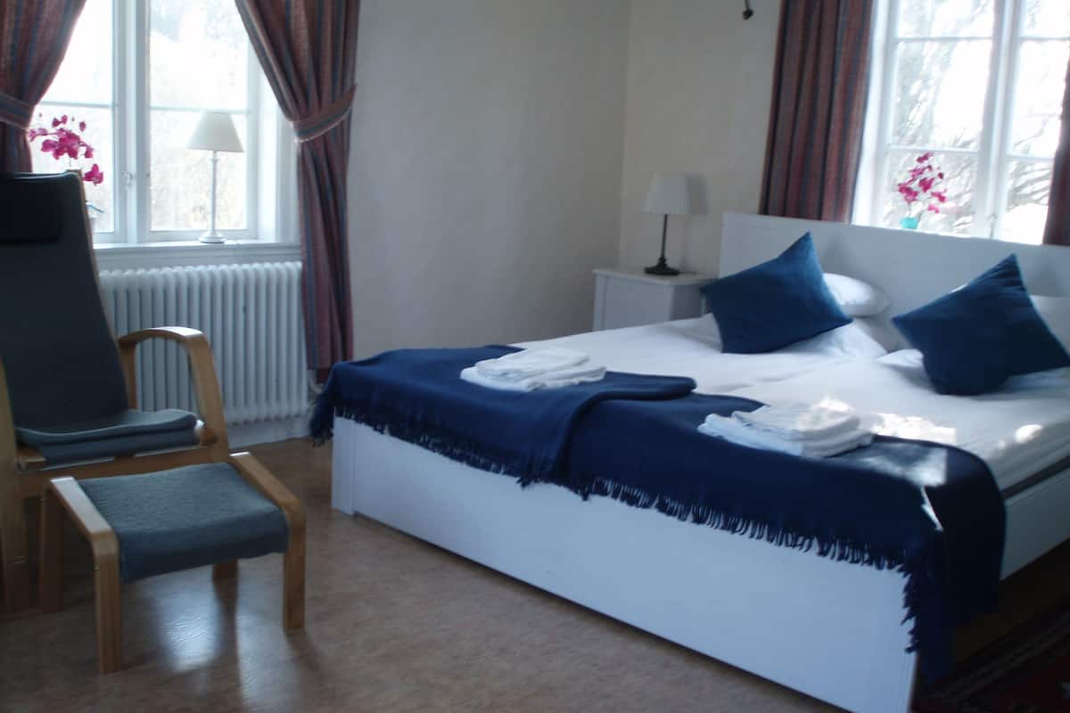 Una cama bien hecha con ropa de cama azul, una silla de madera, una mesita de noche con una lámpara y una ventana con cortinas.