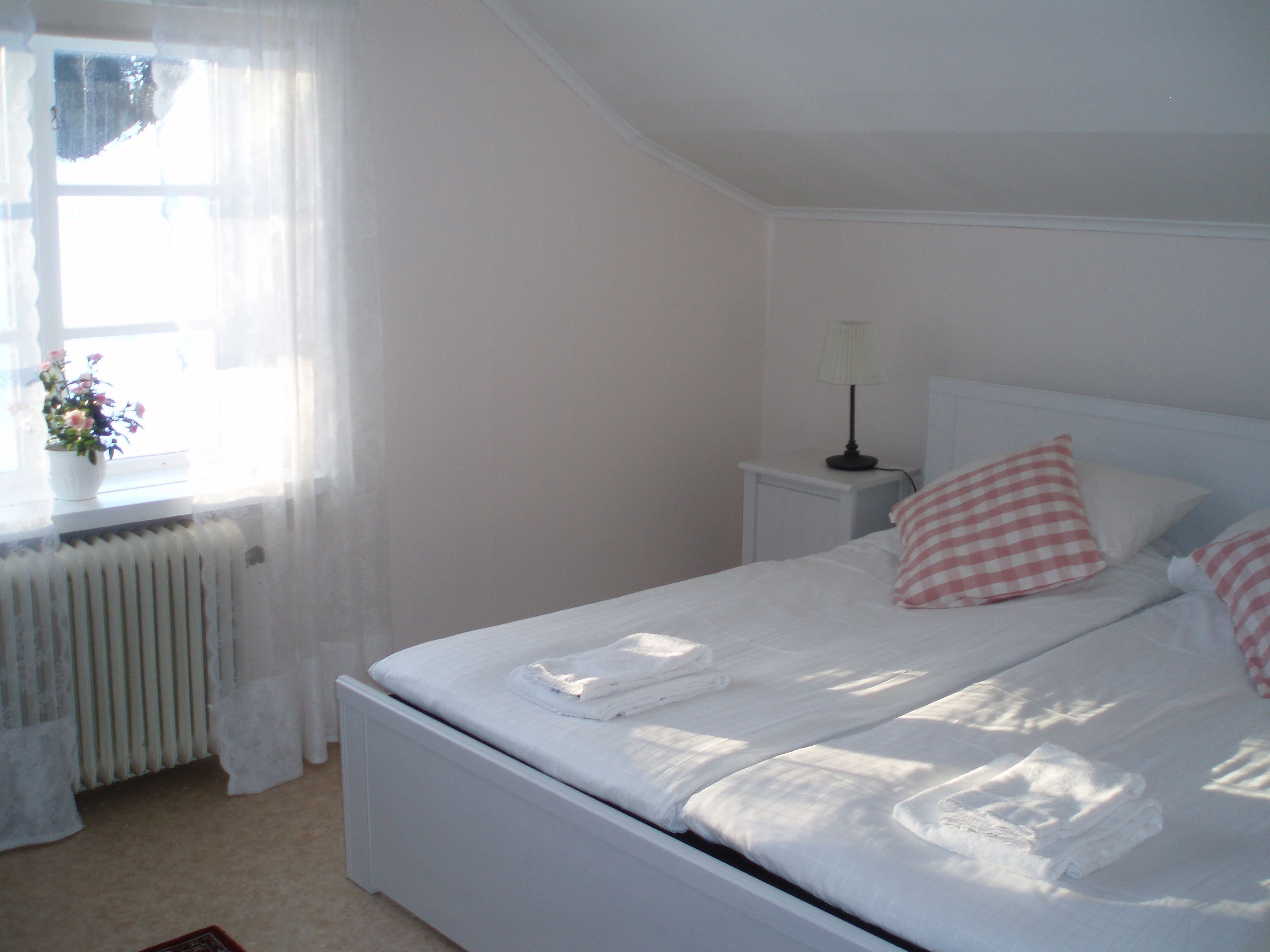 Una cama bien tendida con sábanas blancas y almohadas a cuadros rosas, una mesita de noche con una lámpara y una ventana con cortinas traslúcidas y una planta en maceta.
