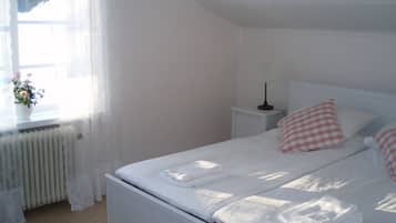 Una cama bien tendida con sábanas blancas y almohadas a cuadros rosas, una mesita de noche con una lámpara y una ventana con cortinas traslúcidas y una planta en maceta.