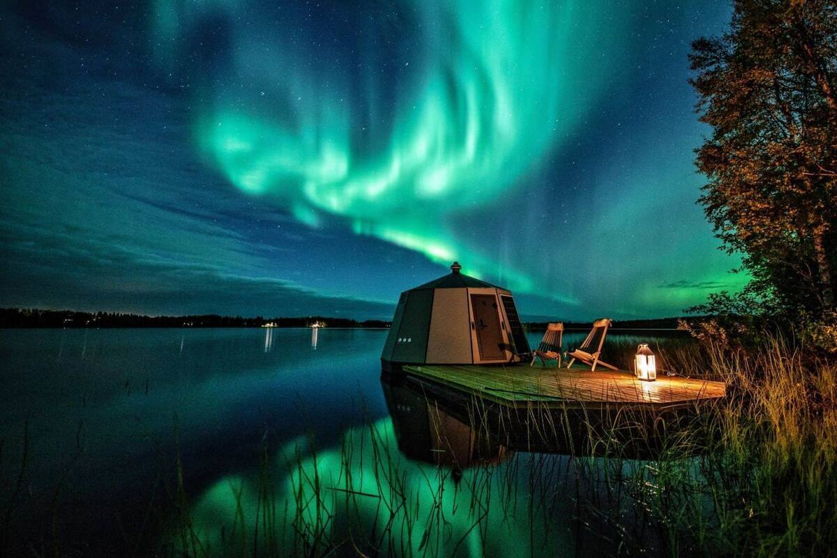 Una cabaña con terraza, dos sillas y un farol junto a un lago tranquilo, bajo una exhibición de auroras boreales.
