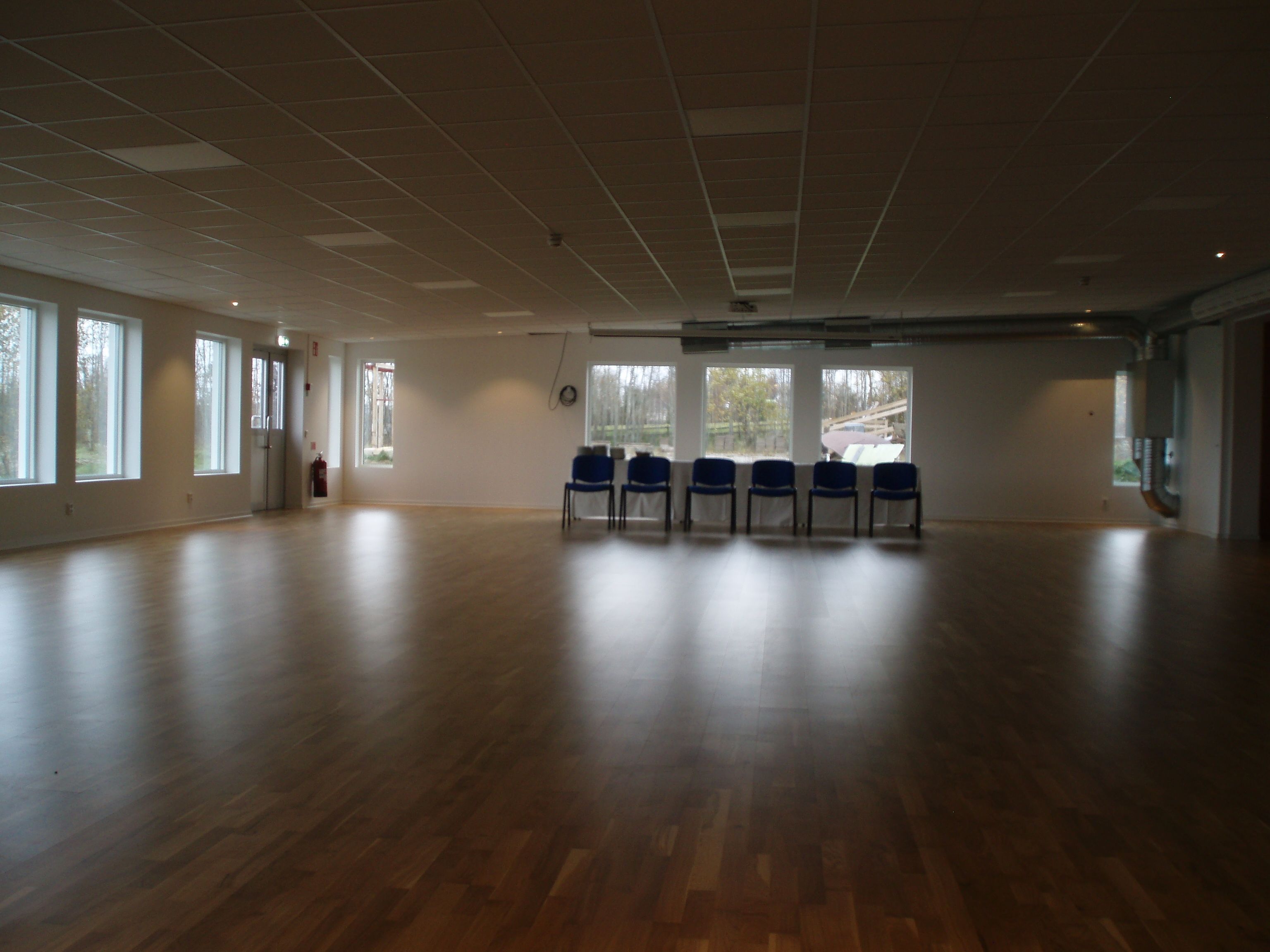 A spacious room with a wooden floor, large windows, and blue chairs arranged in a row.