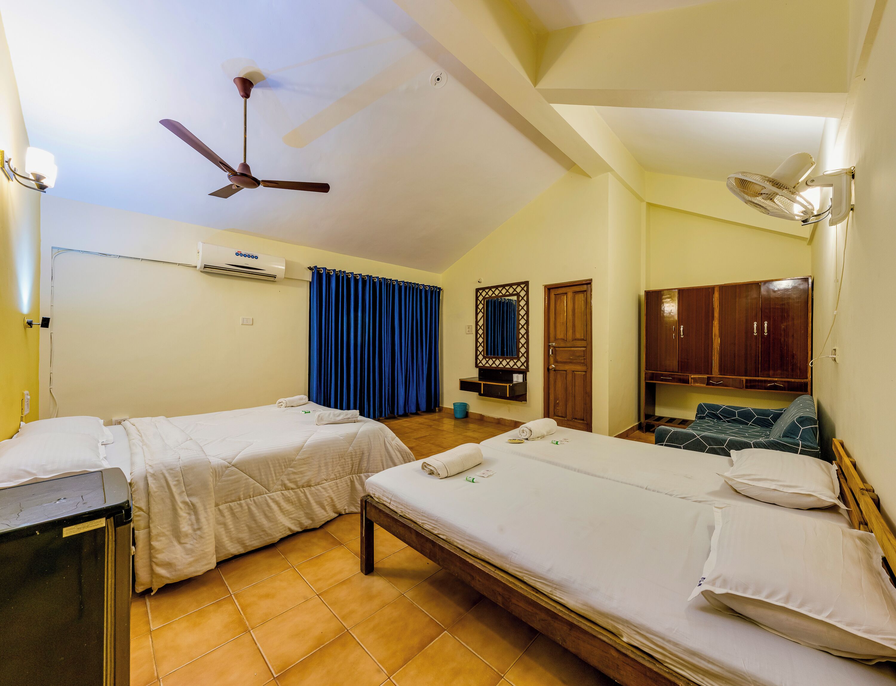 Habitación de hotel con dos camas, ventilador de techo, equipo de aire acondicionado, armario de madera y una mesita con lámpara.