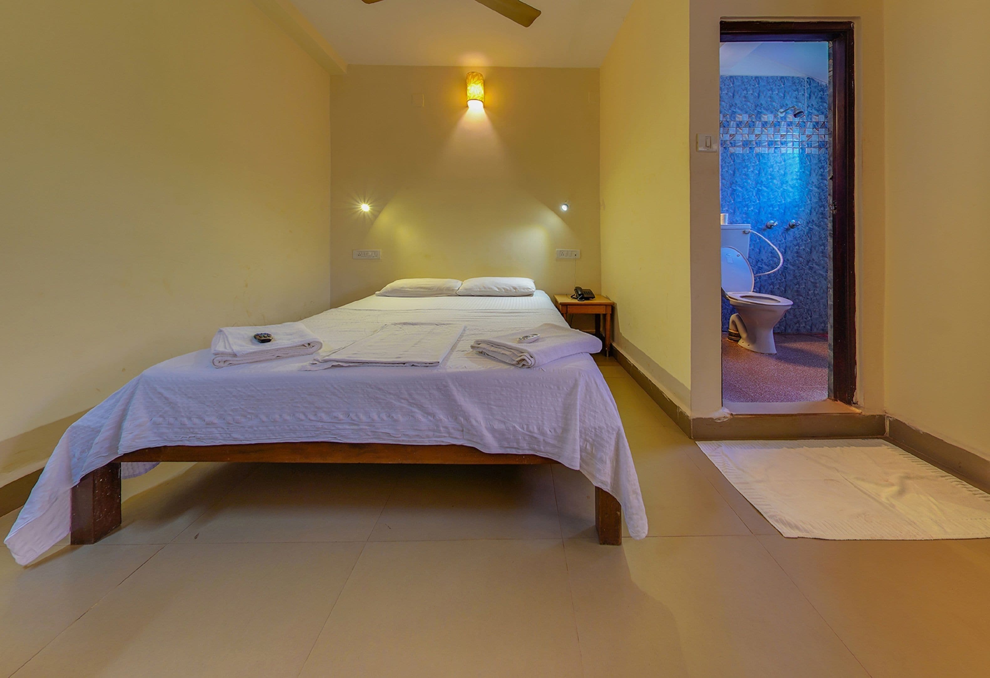 A single bed with white linens, a bedside table with a lamp, and a view of a bathroom with a blue-tiled wall.