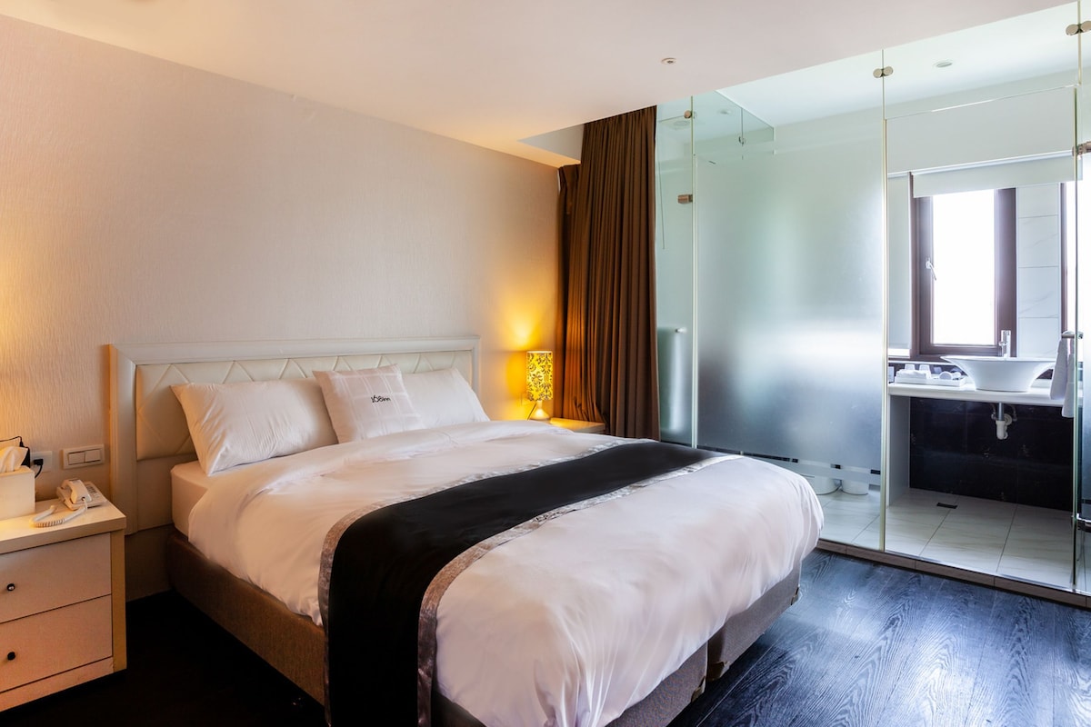A hotel room with a bed, bedside tables, a lamp, a mirror, and a bathroom with a sink and mirror.