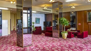 Un hall d’hôtel avec un tapis à motifs, des sièges rouges, un comptoir d’accueil et une grande plante.