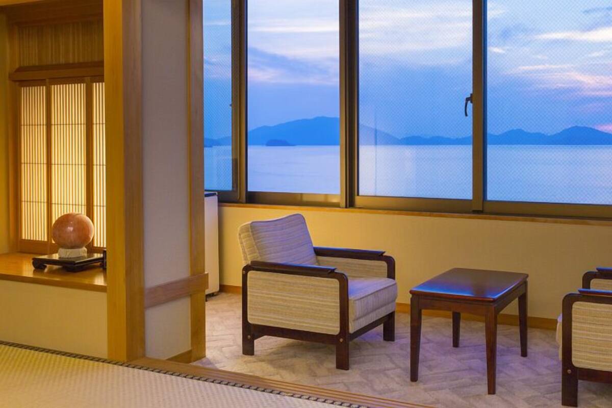 A room with a view of a body of water and mountains, featuring two armchairs and a small table.