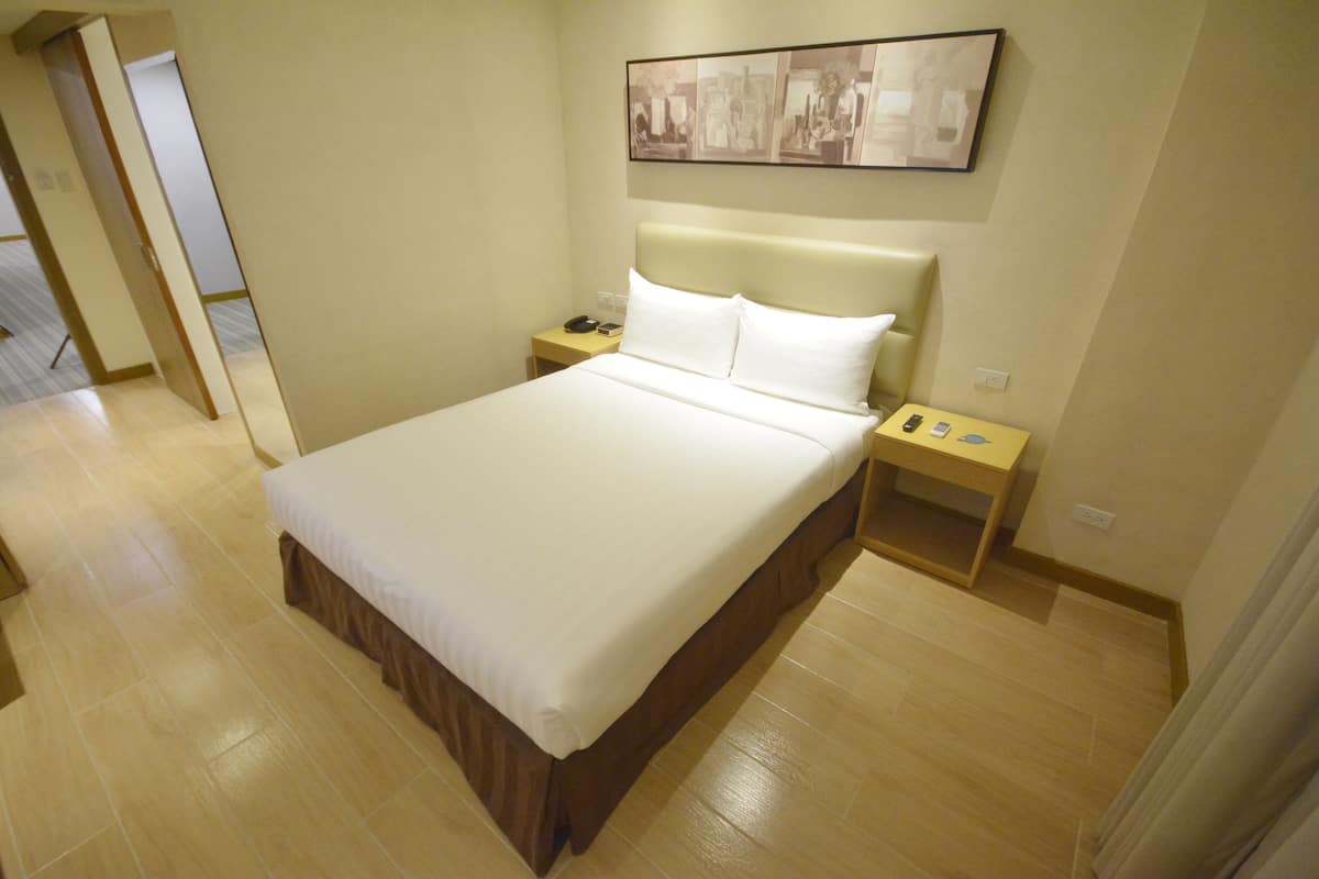Un lit bien fait, avec une tête de lit en bois, une table de chevet sur laquelle se trouvent un téléphone et une petite lampe, et un tableau encadré accroché au mur.