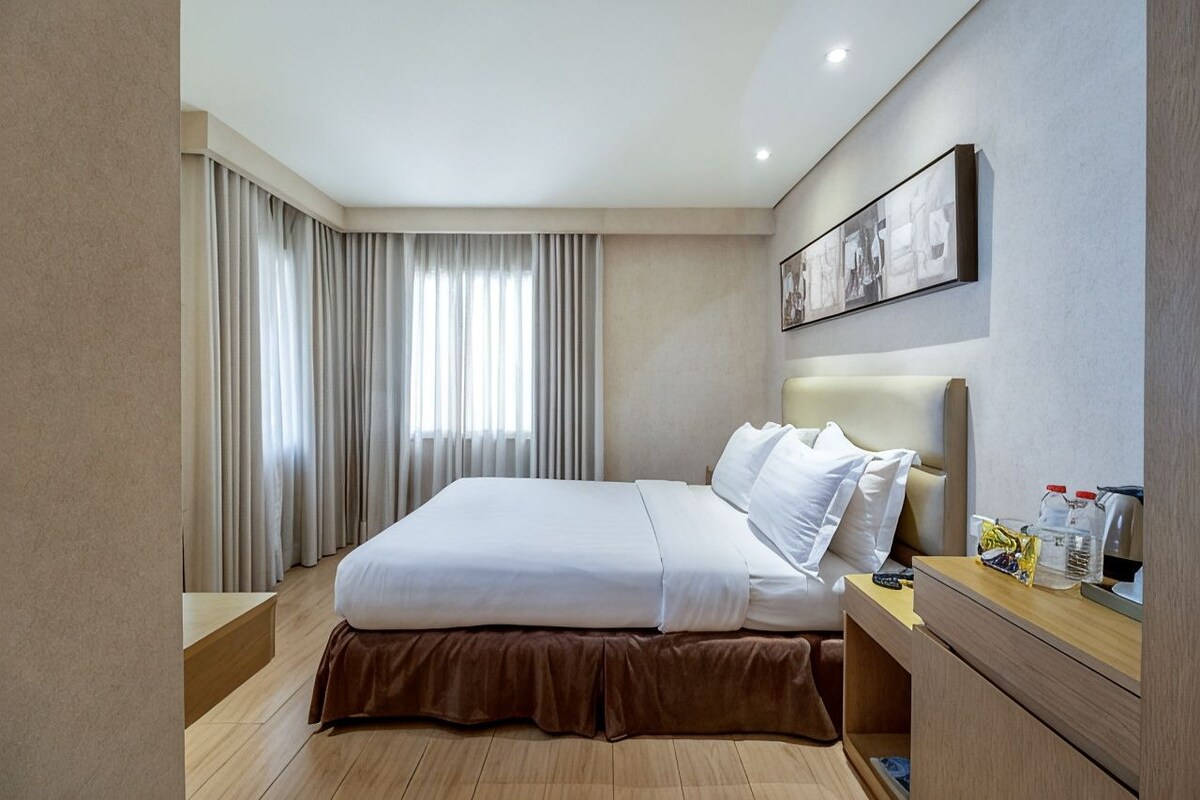 Une chambre d’hôtel comprenant un lit, une table de chevet, une commode, un miroir et une fenêtre avec des rideaux.