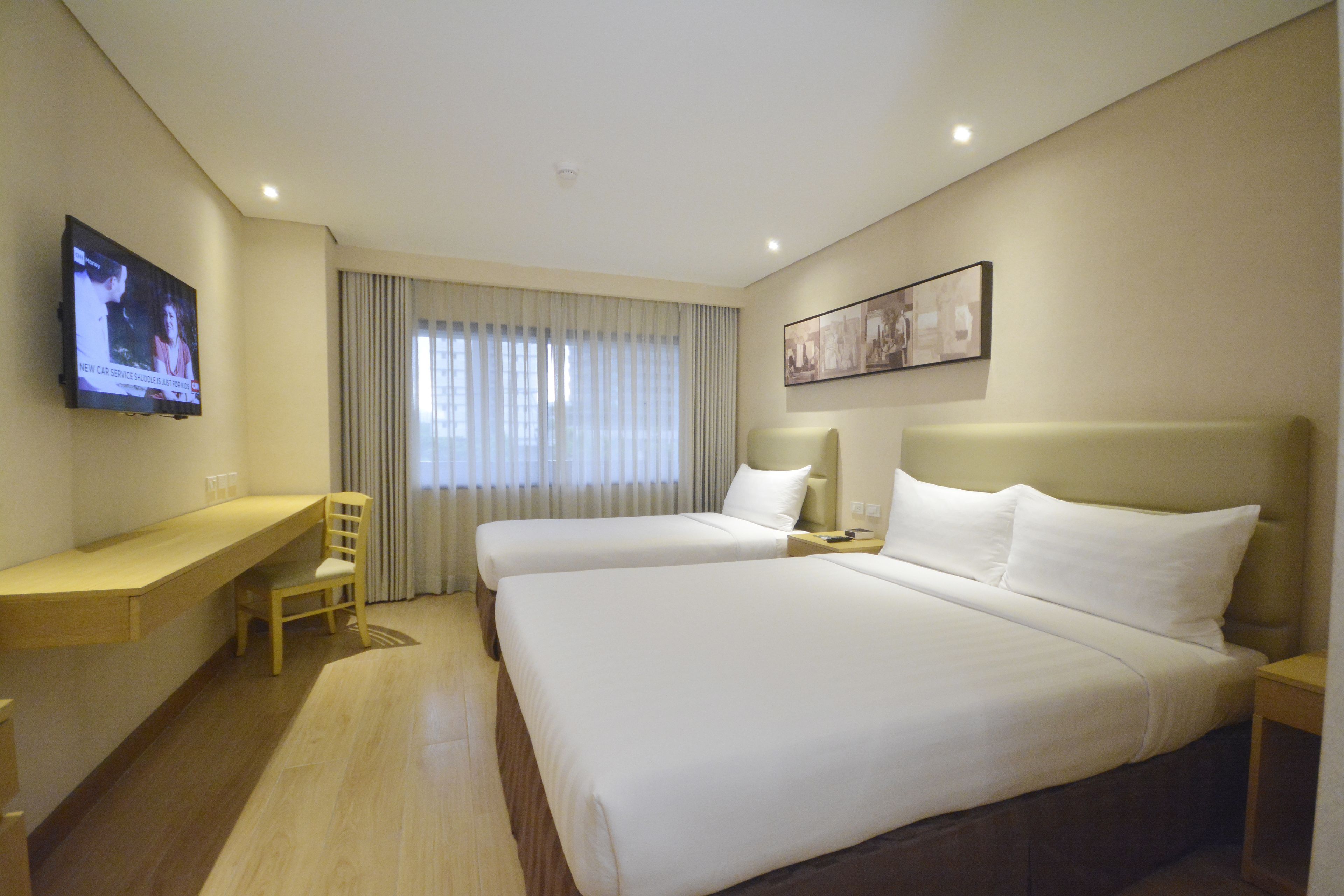 A hotel room with two beds, a desk, a chair, a TV mounted on the wall, and a window with curtains.