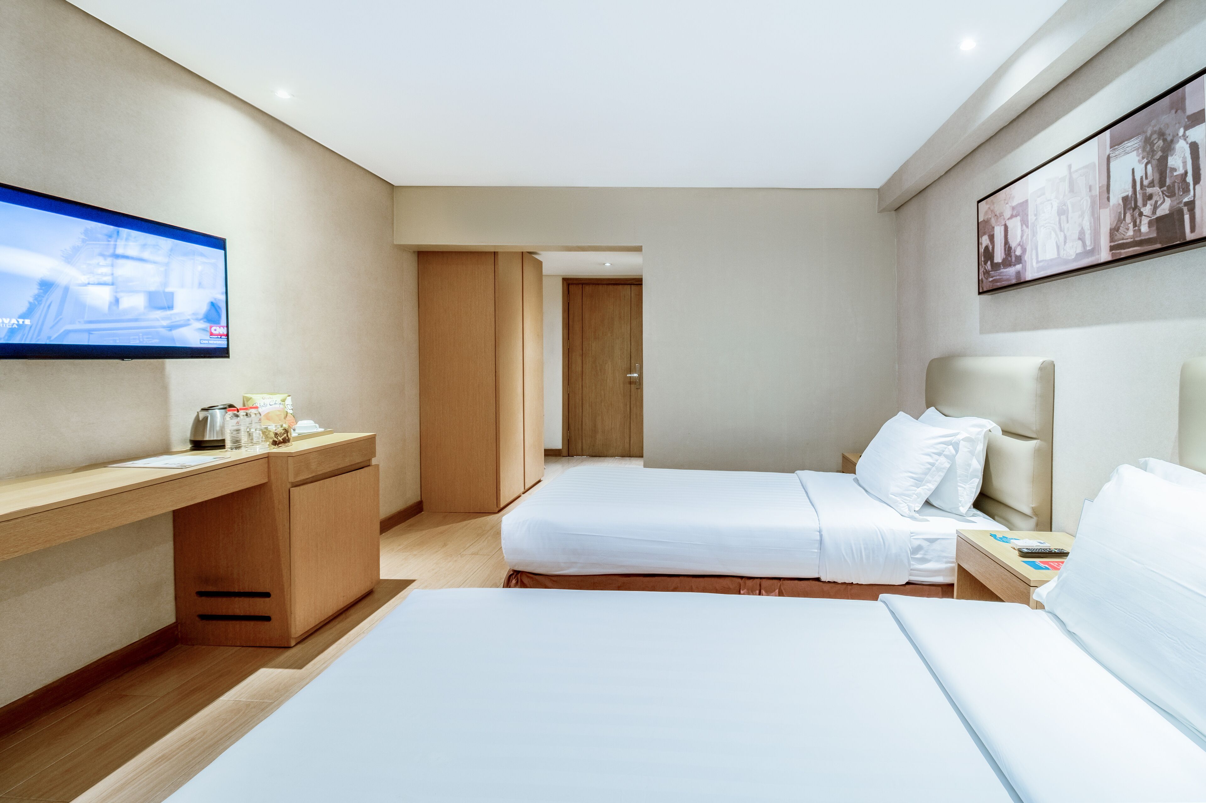 A hotel room with two beds, a desk, a flat-screen TV, and a door leading to another room.