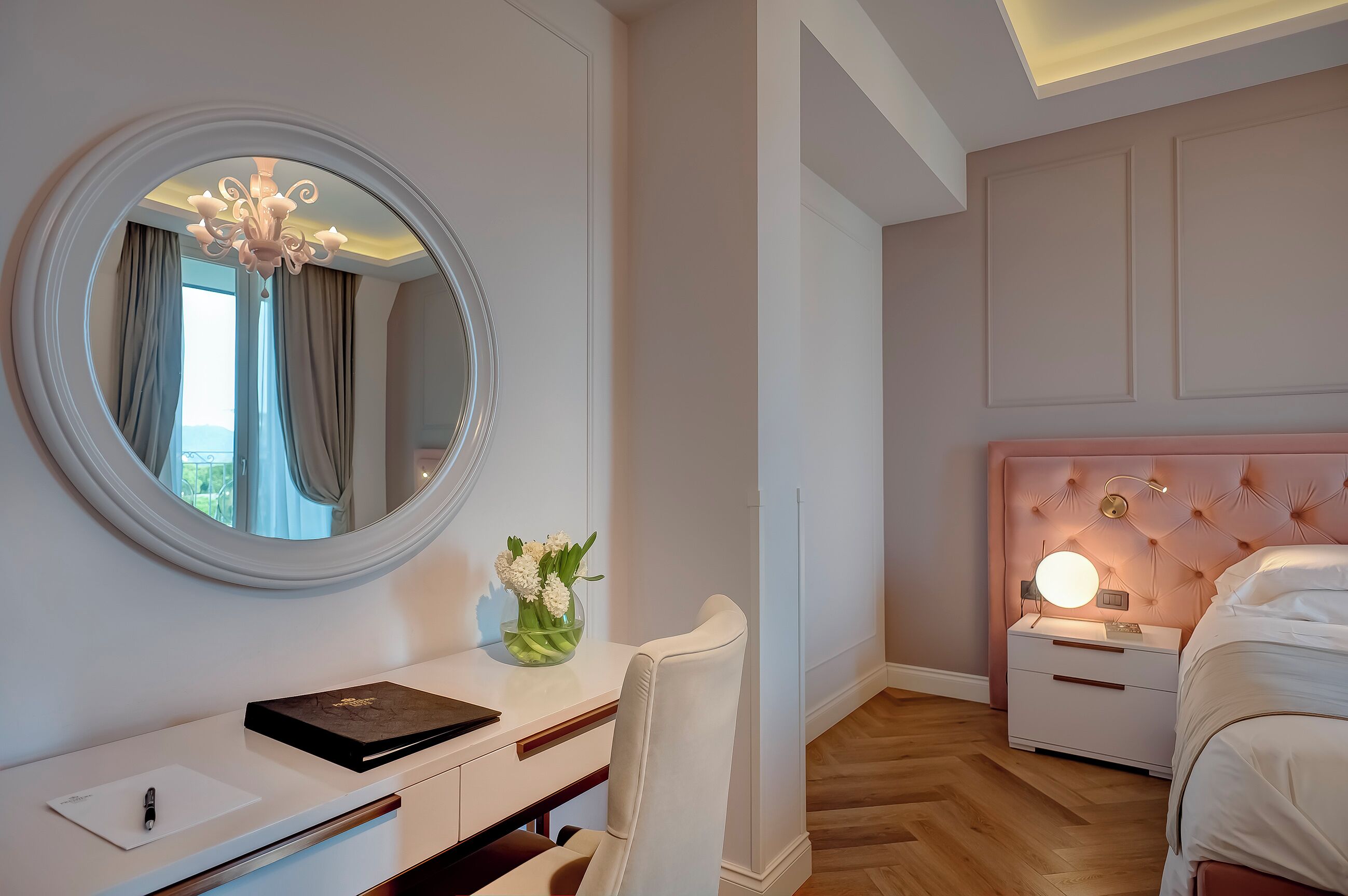 A bedroom with a round mirror, bedside table with a lamp, a bed with a headboard, a desk with a chair, and a vase with flowers.