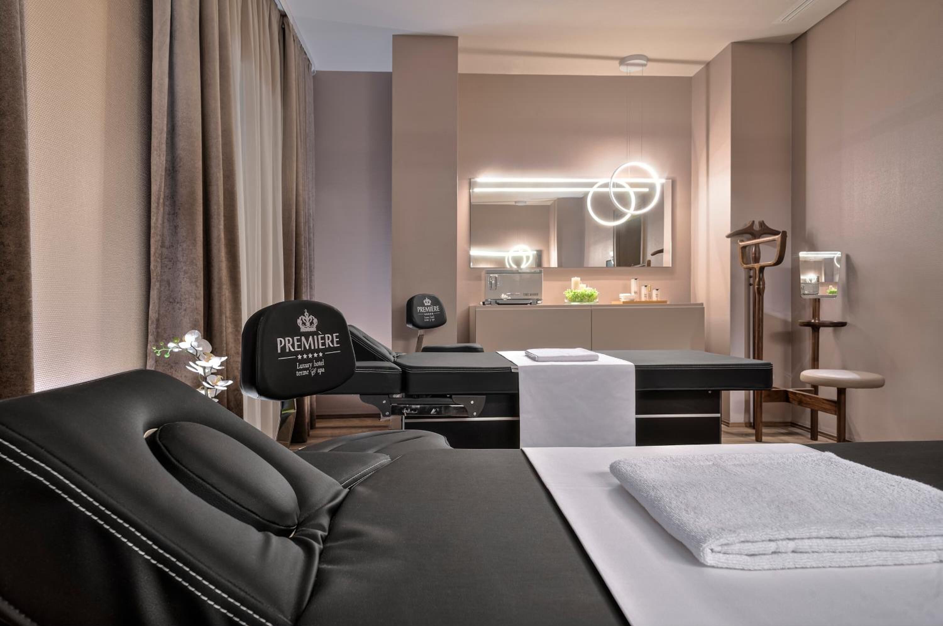 Una camera d'albergo moderna con poltrona massaggiante, scrivania con computer, specchiera e ampia finestra con tende.