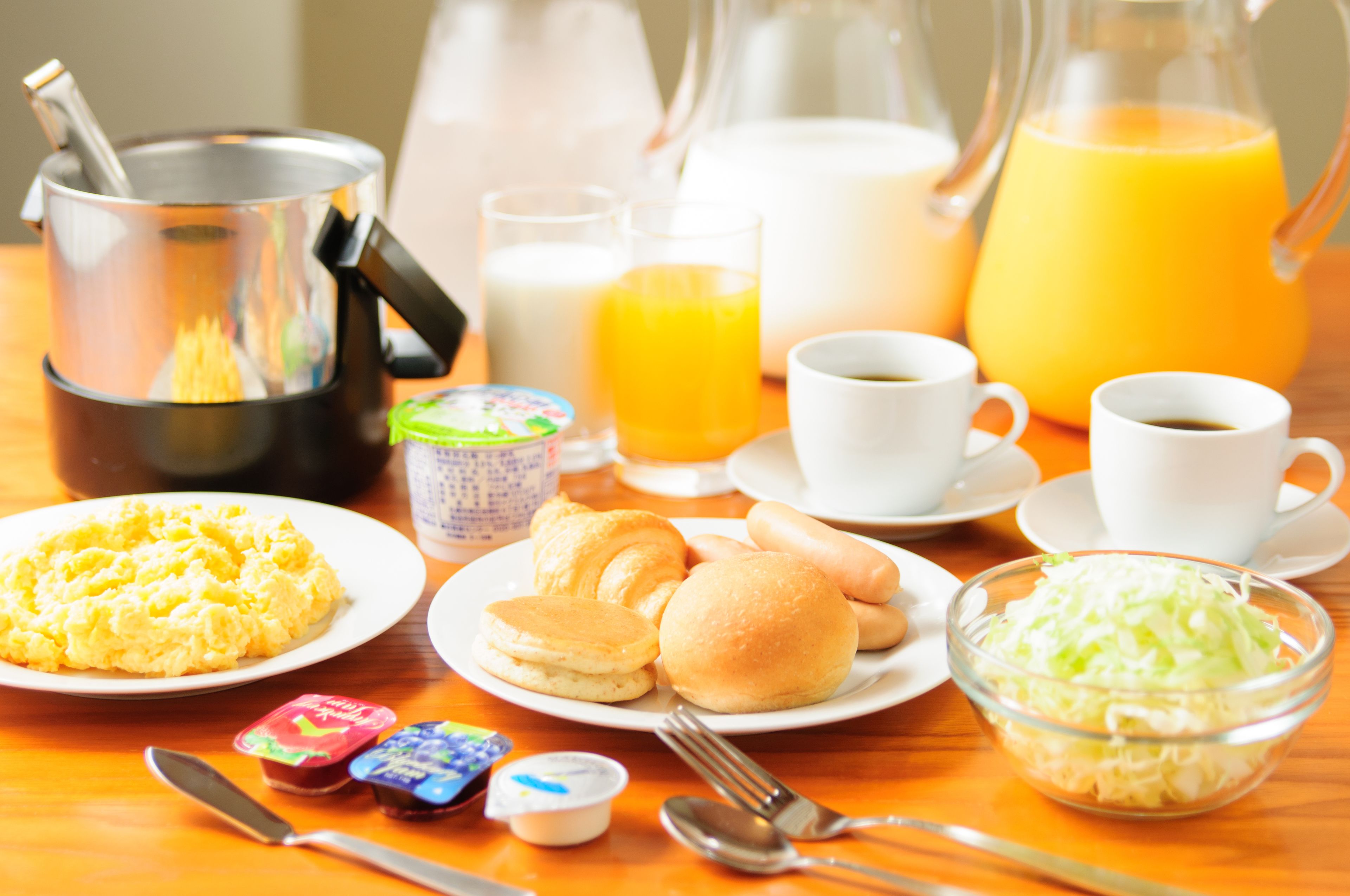 A breakfast setting with various food items, beverages, and utensils on a wooden table.