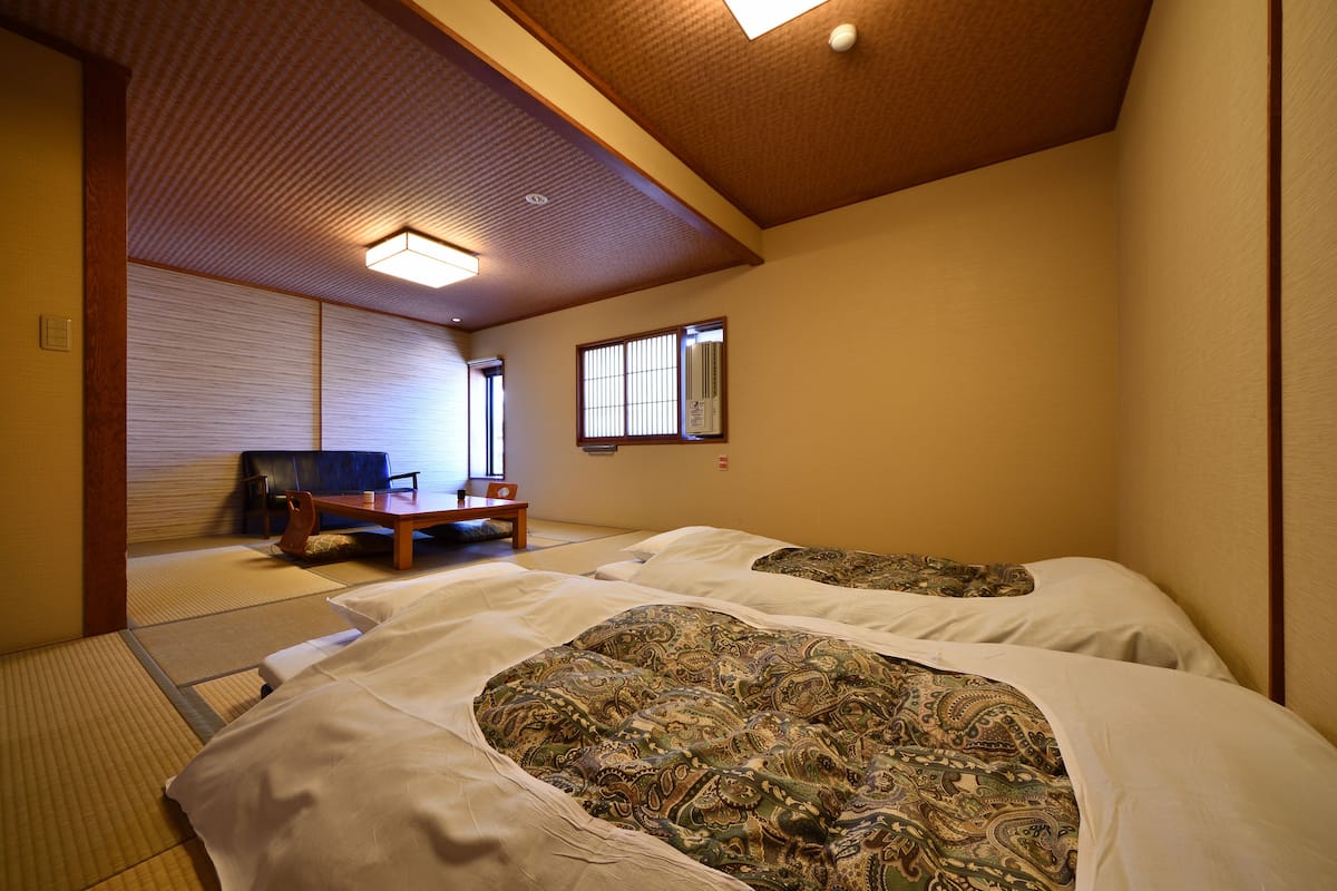 A traditional Japanese-style room with tatami flooring, two beds with patterned bedding, a low wooden table, and a sliding shoji screen door.