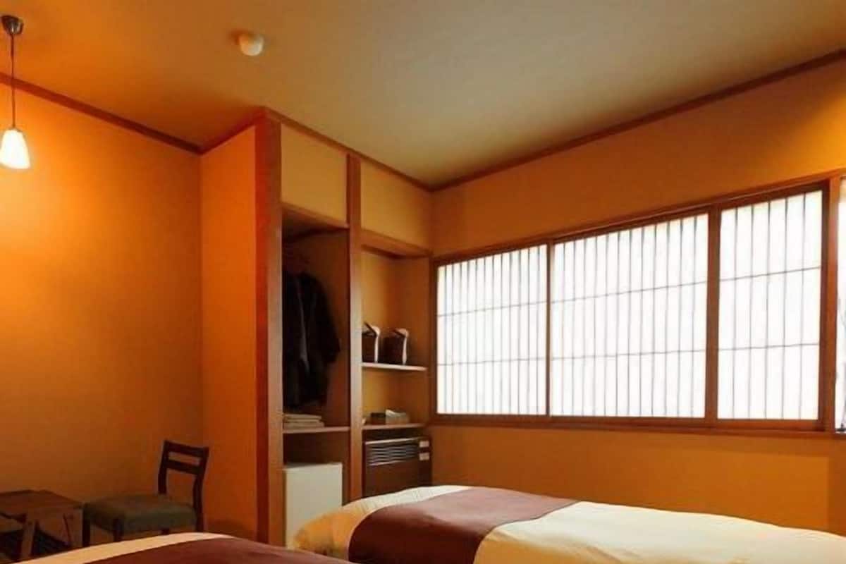 A hotel room with two beds, a wardrobe, a desk, and a window with vertical blinds.