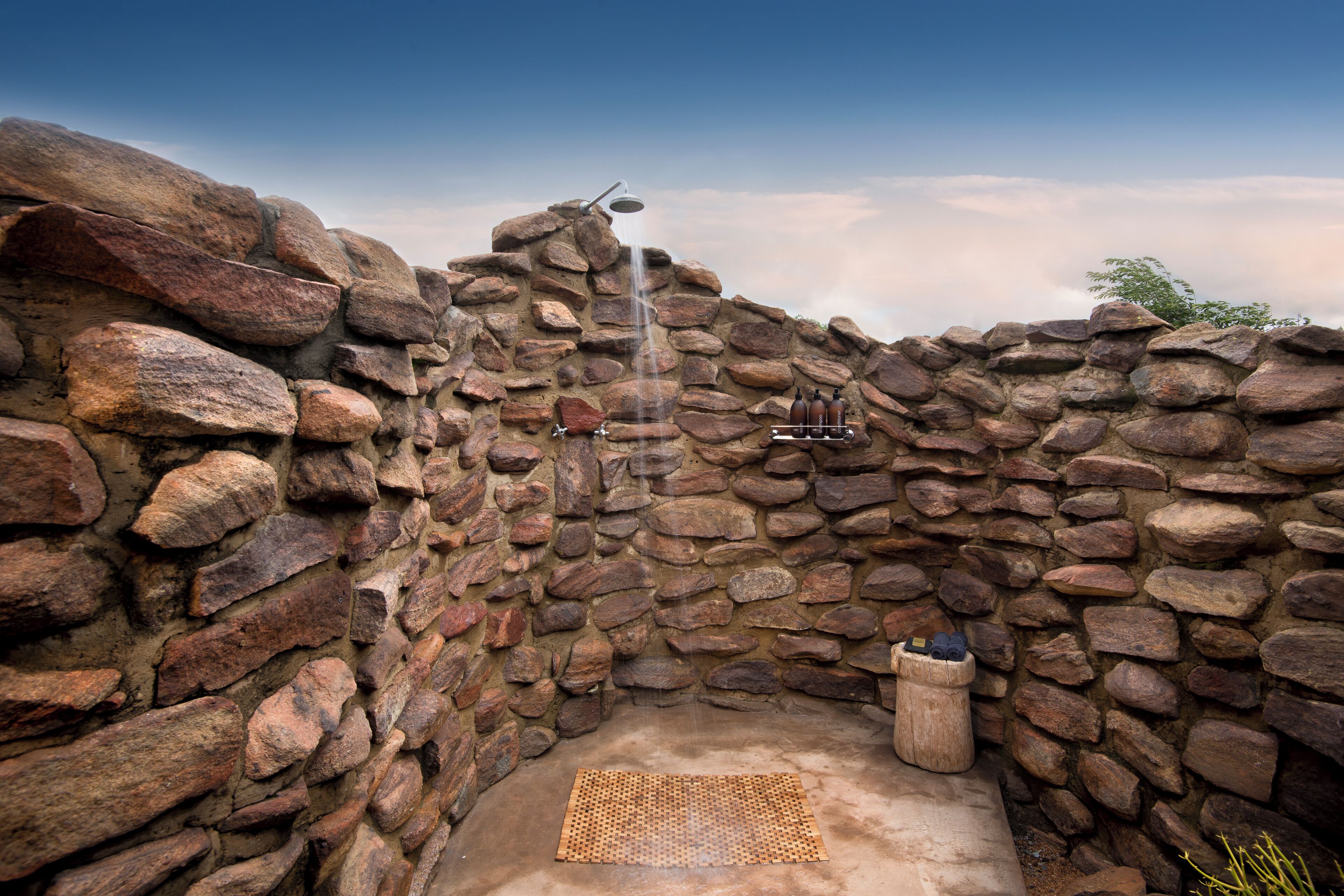 Zona de ducha exterior con paredes de piedra, cabezal de ducha tipo lluvia y un cubo de madera.