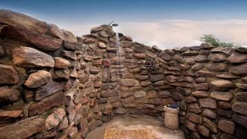 Zona de ducha exterior con paredes de piedra, cabezal de ducha tipo lluvia y un cubo de madera.