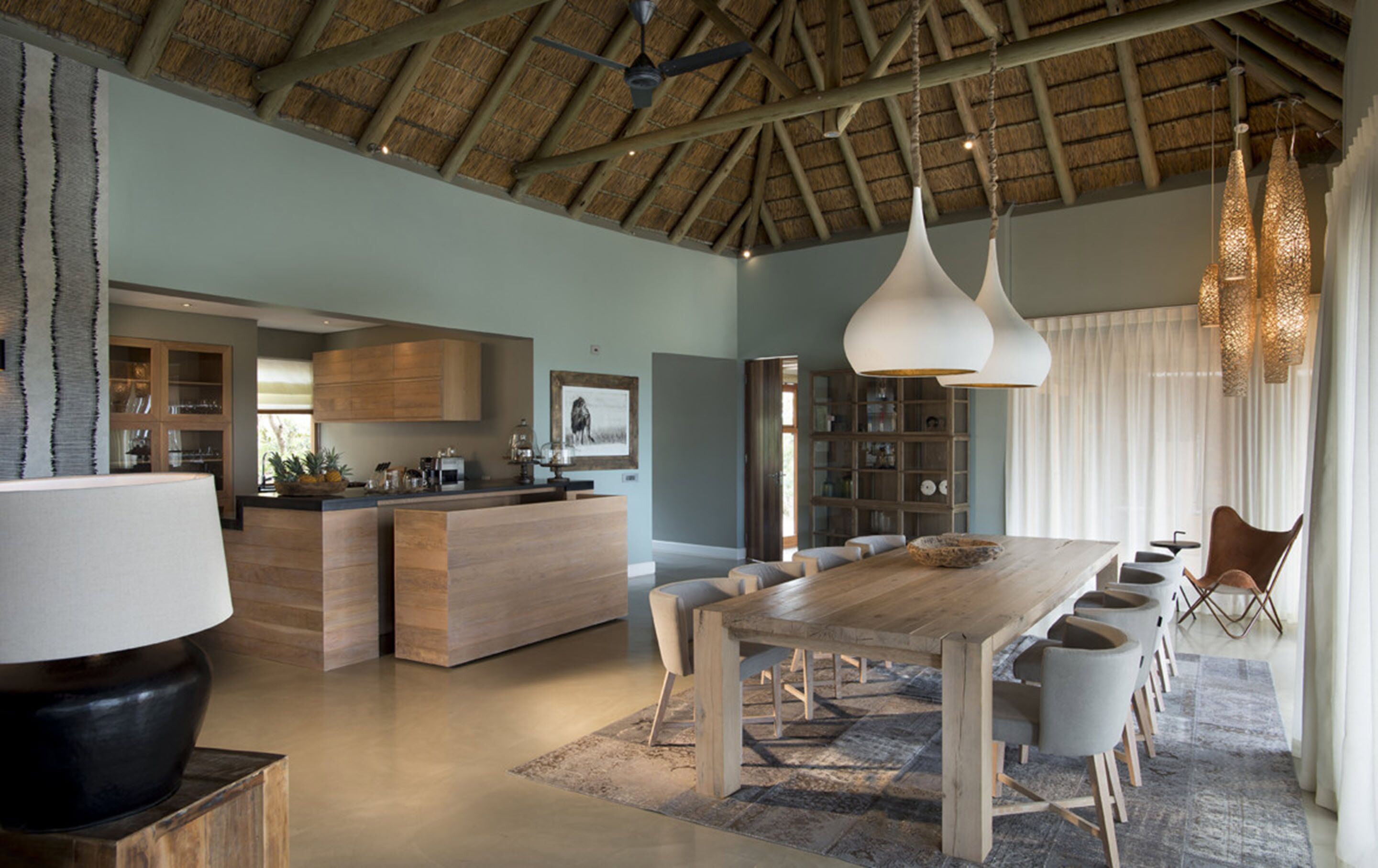 A spacious kitchen with wooden cabinets and a central island, a dining area with a long wooden table and chairs, and a ceiling with exposed beams and hanging lights.