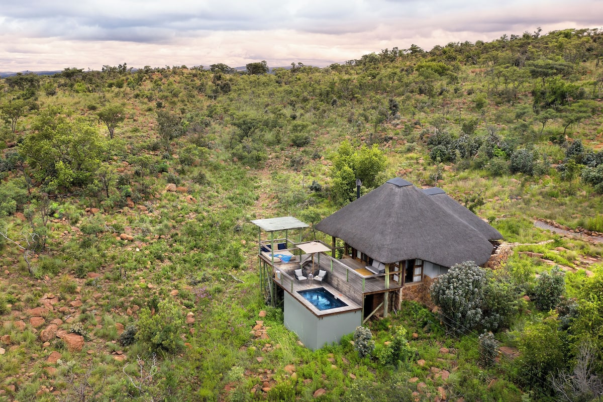 A thatched-roof house with a pool, surrounded by a natural landscape.