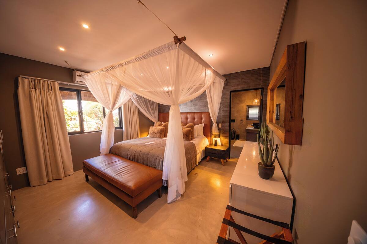 A modern bedroom with a four-poster canopy bed, a leather bench, a bedside table with a lamp, and a potted plant.