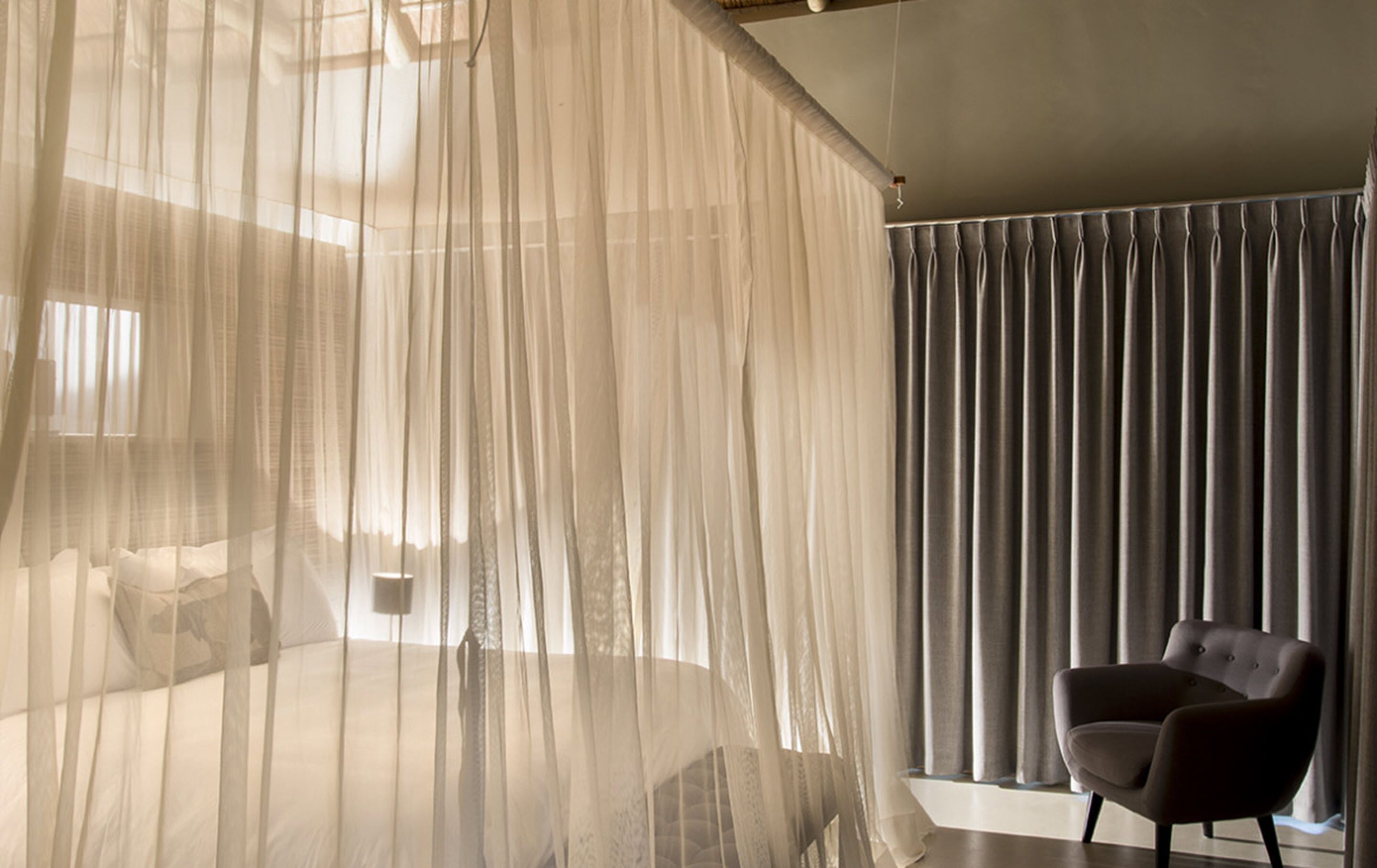 A bedroom with a bed covered by sheer curtains, a bedside table, and a chair by a window with dark curtains.