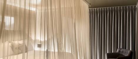 A bedroom with a bed covered by sheer curtains, a bedside table, and a chair by a window with dark curtains.