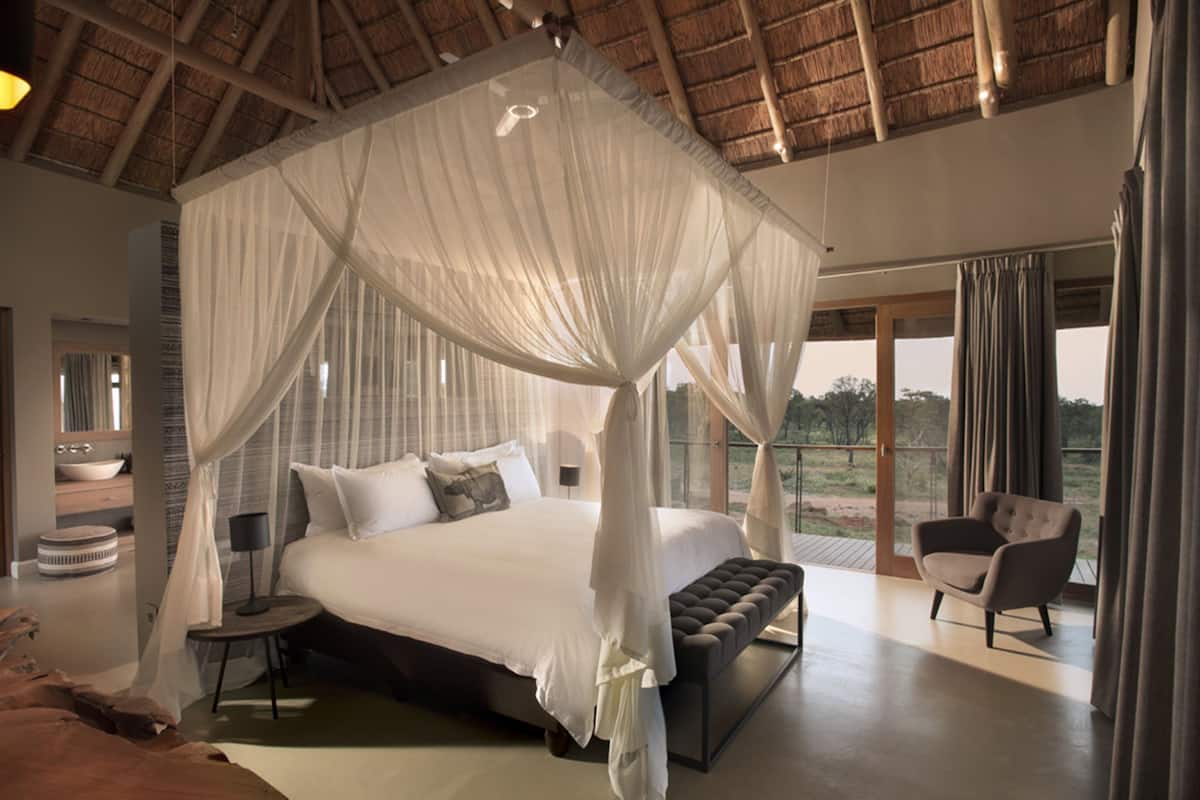 A spacious bedroom with a four-poster bed, a sitting area with a chair, and a large window offering a view of the outdoors.