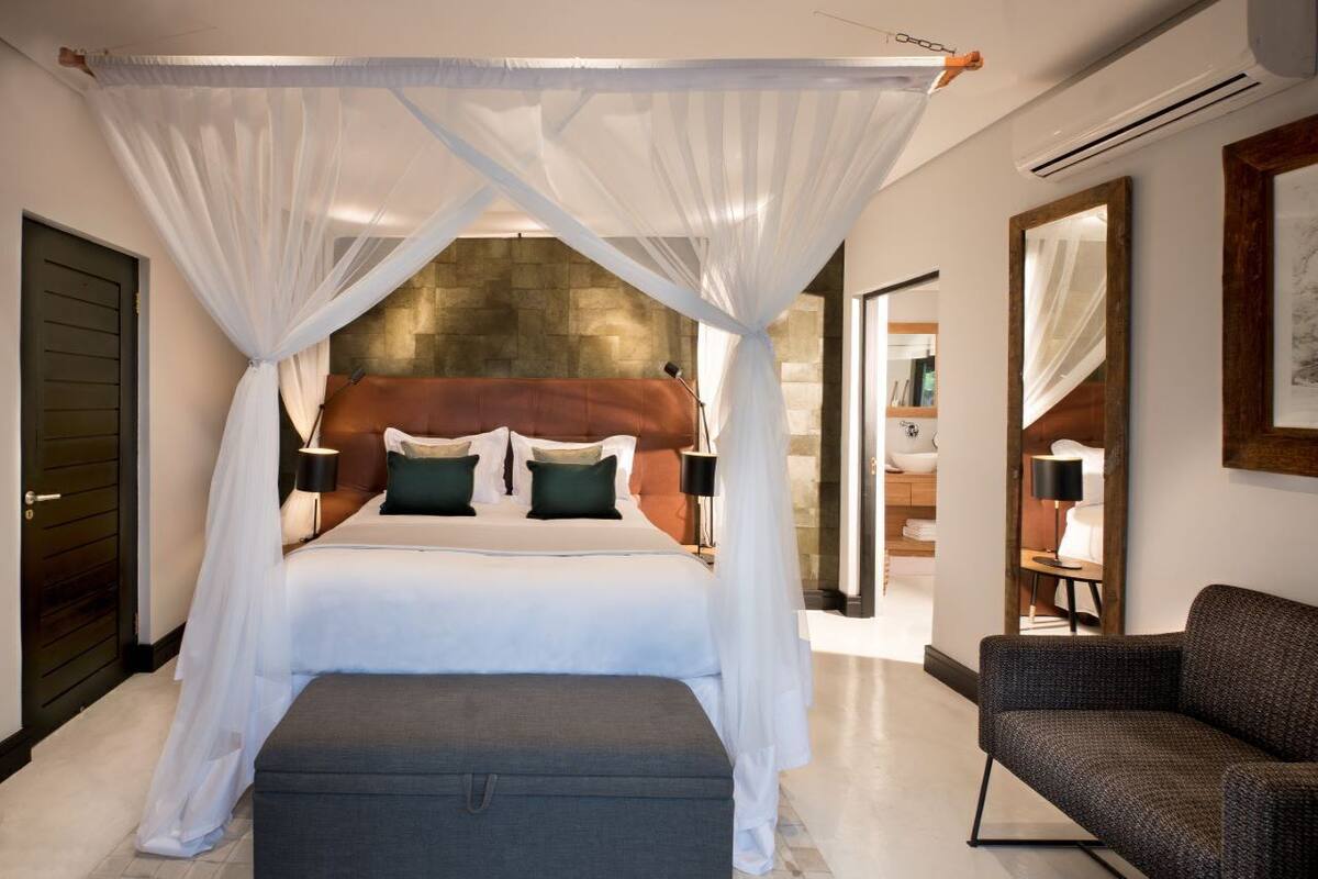 A bedroom with a canopy bed, a sitting area with a chair, and a view of a stone wall.