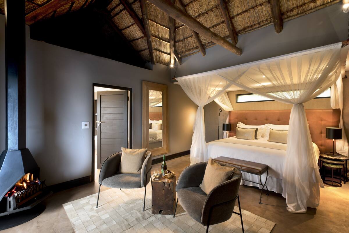 A bedroom with a bed, a bench, two chairs, a fireplace, and a canopy bed.