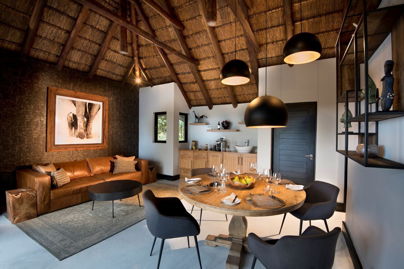 A cozy living room with a round wooden dining table set for six, a leather sofa, and a rustic ceiling.