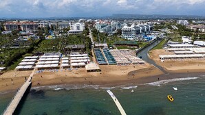 An aerial view of a coastal resort with multiple beach umbrellas, a pier, and a variety of buildings including hotels and resorts. - Side Aluna Hotel (Manavgat)