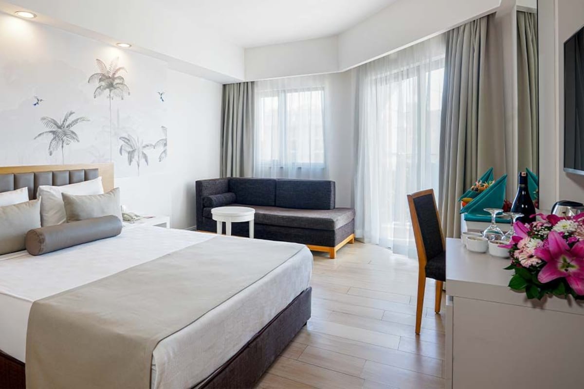 A modern hotel room with a large bed, a sofa, a small table, and a desk with a vase of flowers.