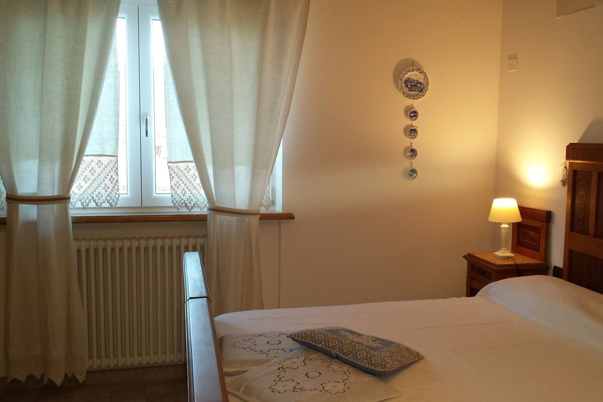 Ein Schlafzimmer mit Bett, Nachttisch mit Lampe, einem Fenster mit Vorhängen, einem Heizkörper und einer Wanddekoration.