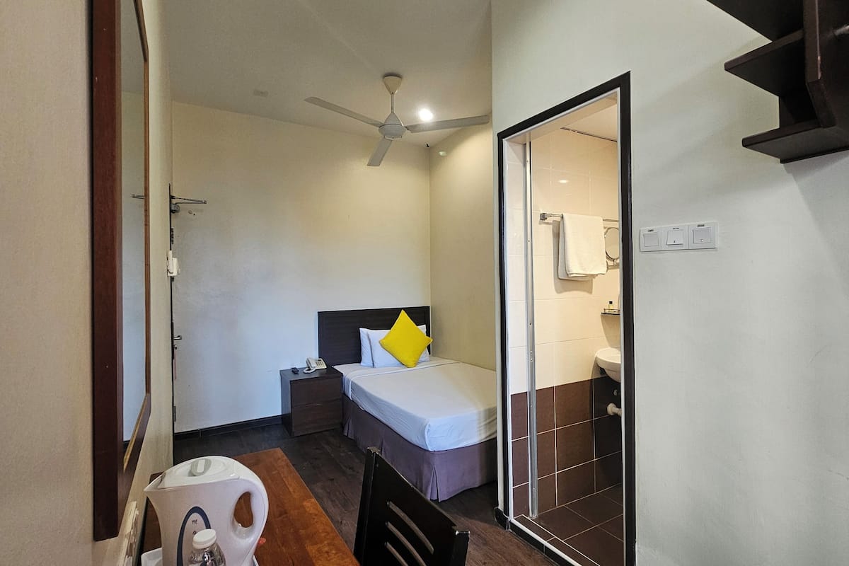 A compact hotel room with a single bed, a bedside table, a nightstand, a ceiling fan, a small table with a kettle, and a bathroom with a toilet and sink.