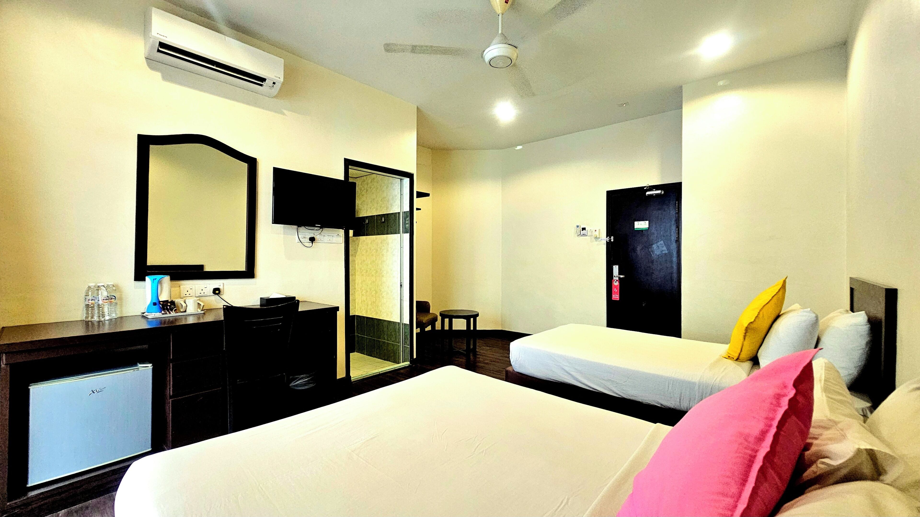 Habitación de hotel con cama, escritorio, silla, espejo, armario, unidad de aire acondicionado, ventilador de techo y puerta con cerradura.