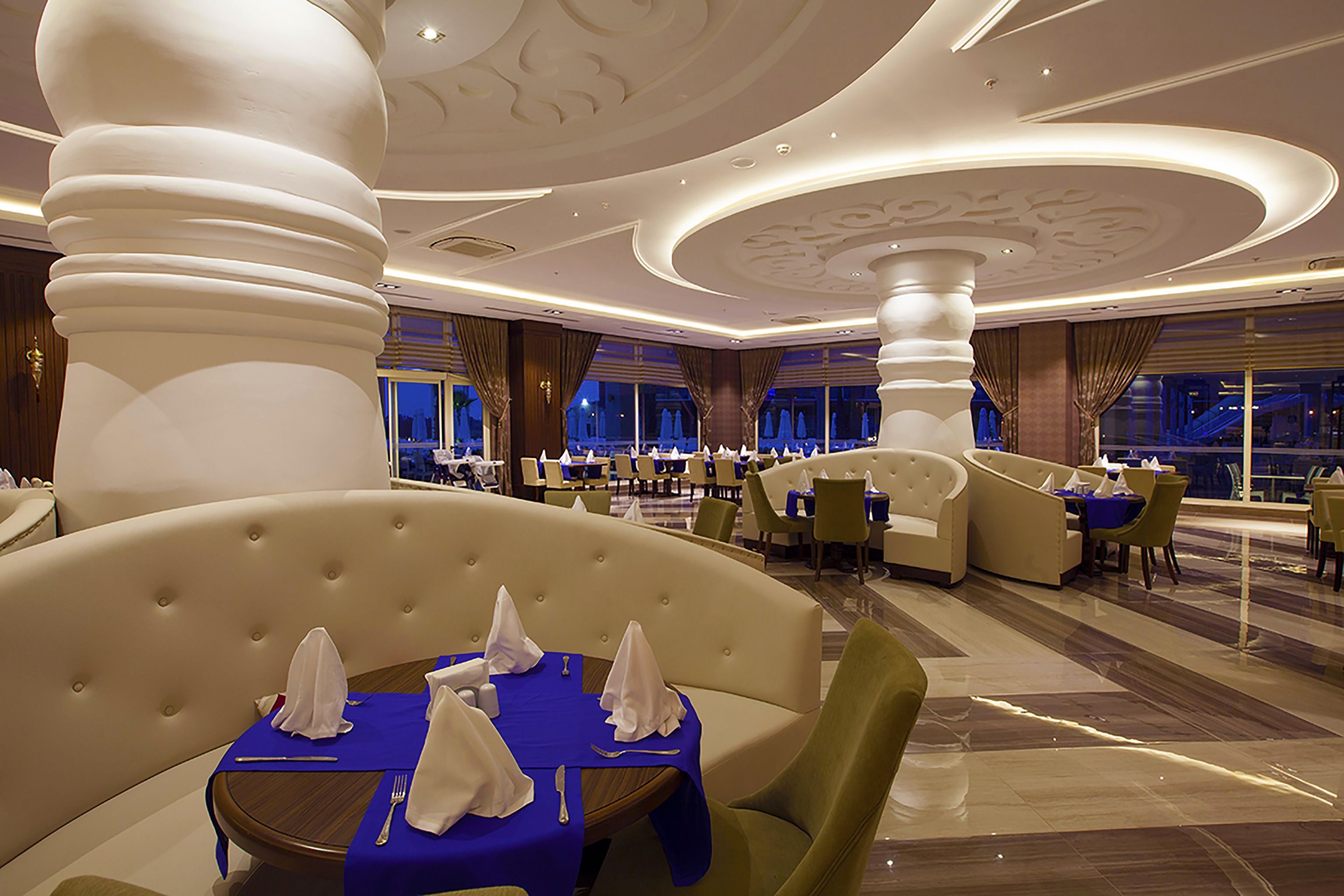 A spacious hotel dining area with multiple tables and chairs, large decorative columns, and a modern ceiling design.