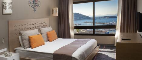 A hotel room with a large bed, a desk, a telephone, and a view of the sea and cityscape.